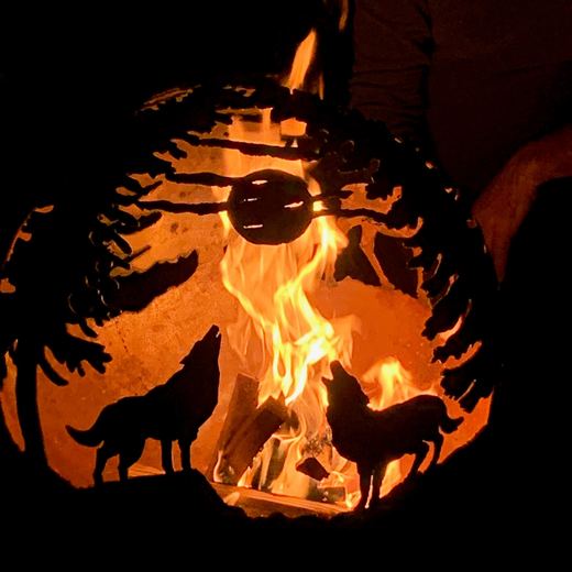 Stahlkugel mit zwei heulenden Wölfen unter Tannen, innen brennt ein Holzfeuer