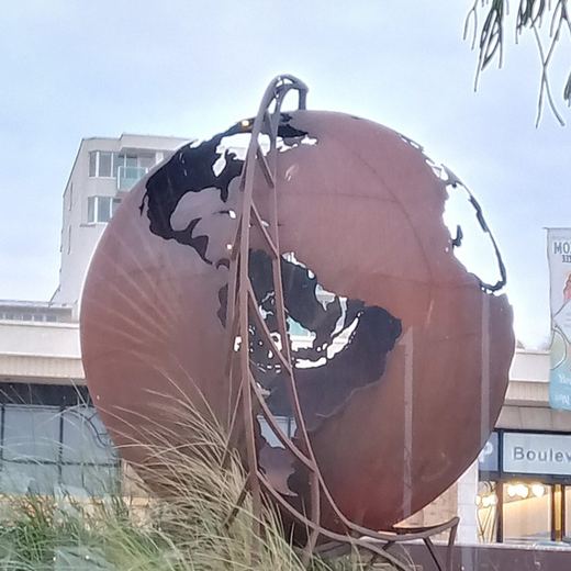 Weltkugel aus rostbraunem Stahl auf ihrem Stahlgestell, aufgestellt im Dünengras an einer Promenade